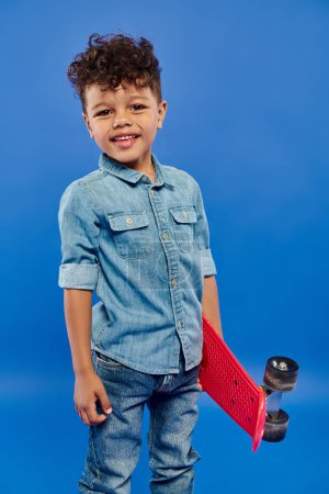 Photo for Happy african american preschooler boy in stylish denim clothes standing on penny board on blue - Royalty Free Image