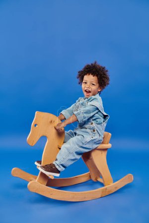 Photo for Happy african american toddler boy in stylish denim clothes sitting on wooden rocking chair on blue - Royalty Free Image