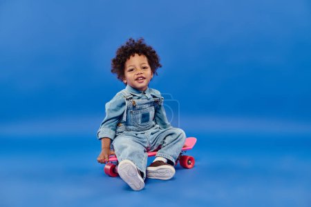 Téléchargez les photos : Joyeux jeune garçon afro-américain en denim assis sur un panneau de penny sur fond bleu - en image libre de droit