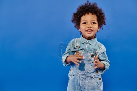 Photo for Adorable and curly african american toddler boy in stylish denim clothes standing on blue background - Royalty Free Image