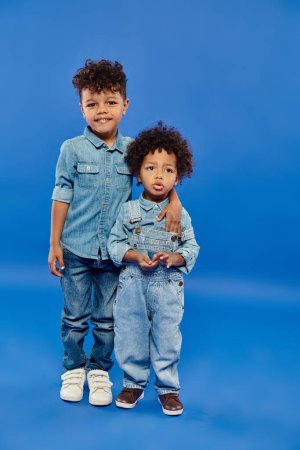 Téléchargez les photos : Heureux afro-américain préscolaire garçon en denim vêtements étreignant petit frère sur fond bleu - en image libre de droit