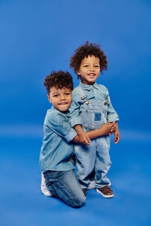Téléchargez les photos : Heureux afro-américain préscolaire garçon en denim vêtements embrassant tout-petit frère sur fond bleu - en image libre de droit