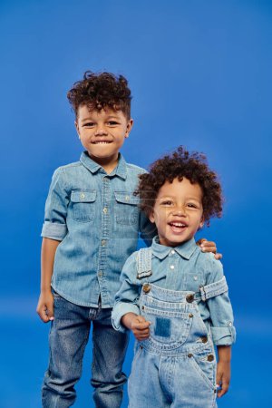 Téléchargez les photos : Joyeux garçon préscolaire afro-américain en vêtements en denim debout avec frère tout-petit sur bleu - en image libre de droit