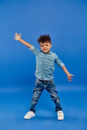 Téléchargez les photos : Joyeux et frisé afro-américain préscolaire garçon en denim vêtements debout et gestuelle sur bleu - en image libre de droit