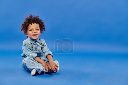 Foto de Alegre y rizado afroamericano niño en ropa de mezclilla sentado con las piernas cruzadas en azul - Imagen libre de derechos