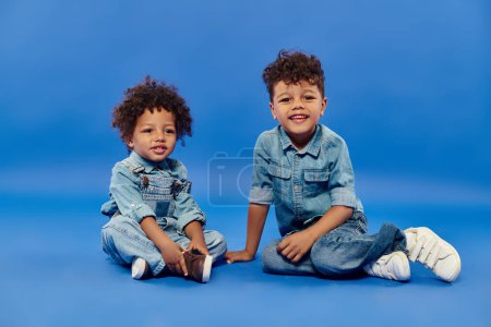 adorables hermanos afroamericanos en ropa de mezclilla con estilo sentados juntos sobre fondo azul