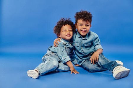 Téléchargez les photos : Adorables frères et sœurs afro-américains en denim élégant assis et étreignant sur fond bleu - en image libre de droit