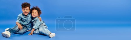 Photo for Adorable african american siblings in stylish denim clothes sitting and hugging on blue, banner - Royalty Free Image