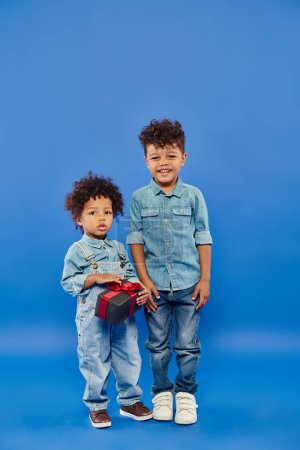 happy african american boy in denim clothes standing near brother with present on blue backdrop