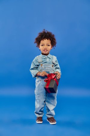 Téléchargez les photos : Heureux afro-américain garçon en denim vêtements debout avec cadeau de Noël sur fond bleu - en image libre de droit