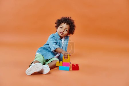 cute african american toddler boy in denim shirt sitting and playing building blocks on orange