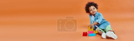 african american toddler boy in denim shirt sitting and playing building blocks on orange, banner