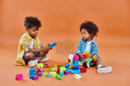 mignon afro-américain frères et s?urs garçon en tenue décontractée assis et jouer blocs de construction sur orange