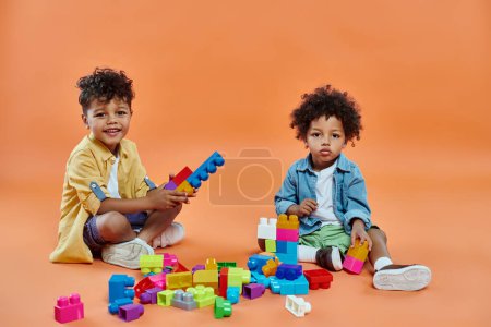 happy african american siblings boy in casual attire sitting and playing building blocks on orange