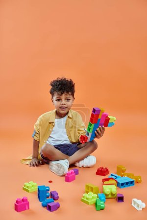 Foto de Niño afroamericano feliz en traje casual sentado y jugando bloques de construcción de colores en naranja - Imagen libre de derechos