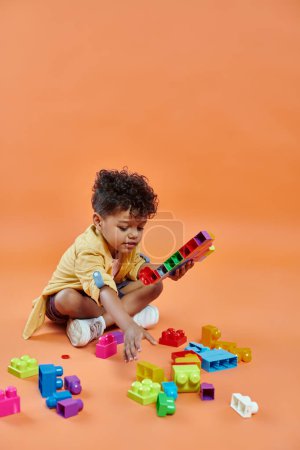 Foto de Niño afroamericano feliz en traje casual sentado y jugando bloques de construcción en fondo naranja - Imagen libre de derechos