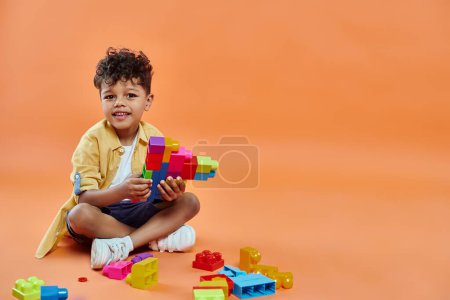 Foto de Alegre afroamericano chico en casual atuendo sentado y jugando bloques de construcción en naranja - Imagen libre de derechos