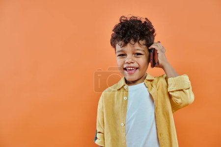 glücklich afrikanisch-amerikanischer Vorschuljunge in lässiger Kleidung, der auf einem Smartphone auf orangefarbenem Hintergrund spricht