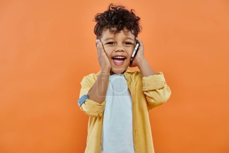 Téléchargez les photos : Joyeux garçon préscolaire afro-américain en tenue décontractée parlant sur smartphone sur fond orange - en image libre de droit