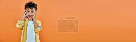 Téléchargez les photos : Heureux garçon préscolaire afro-américain en tenue décontractée parler sur smartphone sur orange, bannière - en image libre de droit