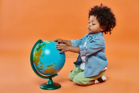 happy african american toddler boy in casual attire touching globe and sitting on orange backdrop