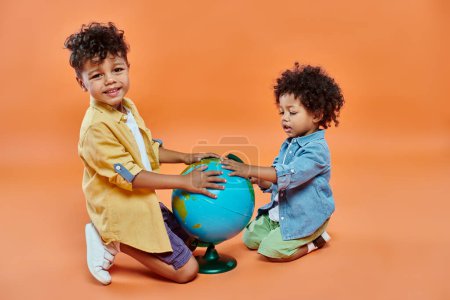happy african american siblings in casual attire touching globe and sitting on orange backdrop