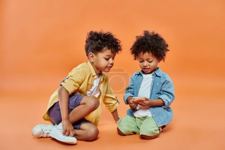 happy african american boy in casual attire looking at toddler brother playing on smartphone