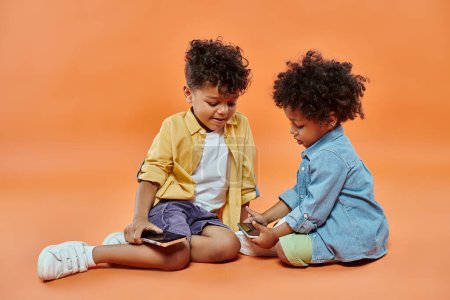 smiling african american boy in casual attire looking at toddler brother playing on smartphone