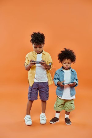 smiling african american siblings in casual summer attire playing on smartphones on orange