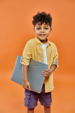 cute and curly african american boy in casual attire smiling and holding laptop on orange background