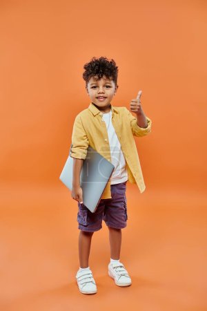 glücklich und lockig afrikanisch amerikanischen Jungen zeigt wie und hält Laptop auf orangefarbenem Hintergrund
