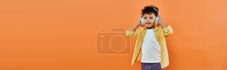 happy and curly african american boy listening music in wireless headphones on orange, banner