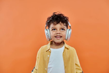 happy african american boy listening music in wireless headphones on orange background, look up