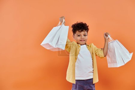 niño afroamericano feliz en traje casual sonriendo y sosteniendo bolsas de compras sobre fondo naranja
