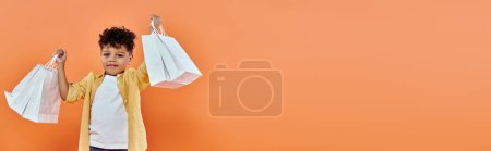 happy african american boy in casual attire holding shopping bags on orange background, banner