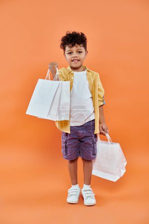 joyful african american boy in casual attire smiling and holding shopping bags on orange background