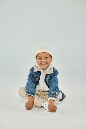 Téléchargez les photos : Heureux afro-américain préscolaire garçon en tenue d'hiver et bonnet chapeau assis sur fond gris - en image libre de droit