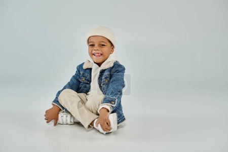 joyful african american preschooler boy in winter attire and beanie hat sitting on grey background