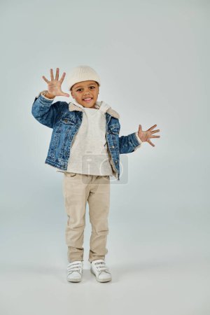 cheerful african american preschooler boy in winter attire and beanie hat gesturing on grey backdrop