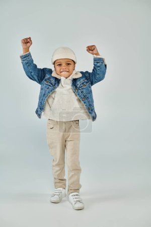excited african american preschooler boy in winter attire and beanie hat gesturing on grey backdrop
