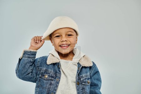 happy african american preschooler boy in winter attire pulling beanie hat on grey backdrop