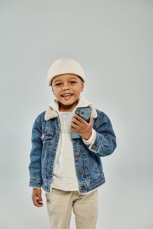 niño preescolar afroamericano feliz en traje de invierno y sombrero de gorro usando teléfono inteligente en gris