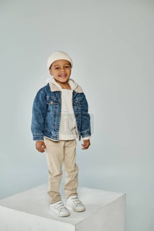 cheerful african american preschooler boy in winter attire and beanie hat on grey backdrop