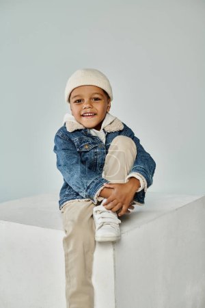 happy african american boy in winter attire and beanie hat sitting on concrete cube on grey