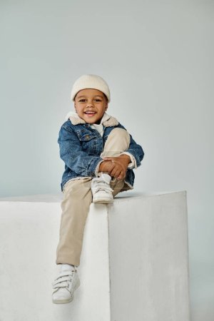 cute happy african american boy in winter attire and beanie hat sitting on concrete cube on grey