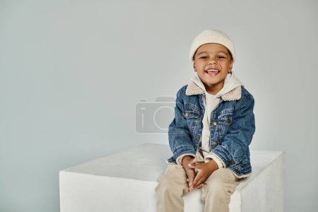 cute joyful african american boy in winter attire and beanie hat sitting on concrete cube on grey