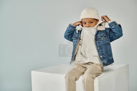 cute african american boy in winter attire and beanie sitting on concrete cube and adjusting hood