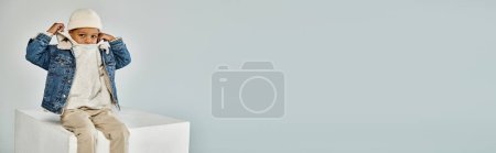 Photo for Banner, african american boy in winter attire and beanie sitting on concrete cube and adjusting hood - Royalty Free Image