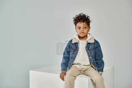 curly african american boy in winter attire and beanie sitting on concrete cube on grey backdrop