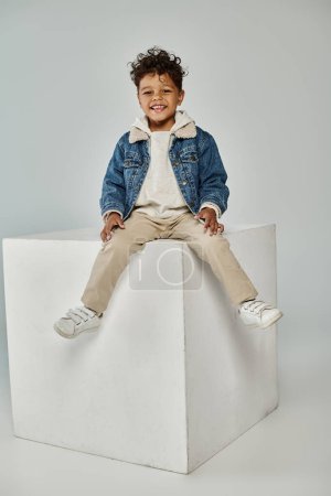 happy african american boy in winter attire and beanie sitting on concrete cube on grey backdrop